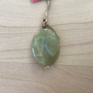 May include: A green oval pendant with a gold wire wrap and a gold bail. The pendant has the letter "K" etched into it.