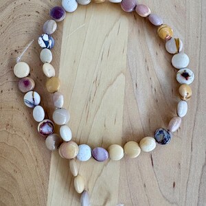 May include: A necklace made of smooth, oval-shaped stones in various colors, including white, beige, purple, and brown. The stones are strung together on a thin, clear cord, creating a natural and earthy aesthetic.