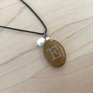 May include: A brown oval pendant necklace with the letter "E" engraved on it. The necklace has a black cord and a white pearl charm.