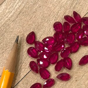May include: A collection of faceted, pear-shaped, deep red gemstones scattered on a light brown wooden surface. A yellow and wood pencil is in the image for scale. The gemstones have a brilliant cut, reflecting light.