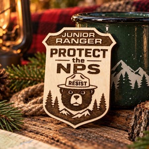 May include: A light-colored wooden badge shaped like a shield featuring laser-engraved text that reads JUNIOR RANGER PROTECT the NPS and an illustration of a bear wearing a hat labeled RESIST, surrounded by pine trees and mountain silhouettes.