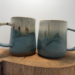 May include: Two ceramic mugs featuring a two-tone glaze with a creamy white top that transitions into a dusty blue base. The mugs have a smooth, rounded body and a curved handle on the side, showcasing a reactive glaze effect with brown drips.