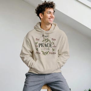 May include: A beige hooded sweatshirt featuring a central graphic with the word PEACE in large letters, surrounded by olive branch illustrations and the word peace translated into multiple languages including Spanish, French, Italian, and German.