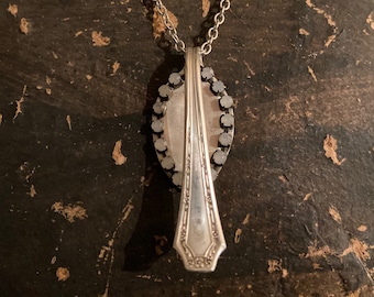 May include: A silver spoon necklace with a decorative design on the handle. The spoon is attached to a silver chain and has a circular design with rhinestones around the edge.
