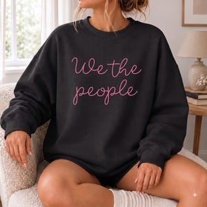 May include: A black long-sleeve crewneck sweatshirt featuring the text "We the people" written in a light pink cursive script font across the center of the chest. The garment has a ribbed neckline, cuffs, and hem, with a relaxed fit.