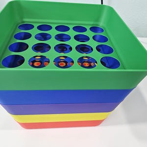 Puede incluir: Un recipiente de plástico cuadrado de varios niveles con una sección superior de color verde brillante que presenta una rejilla de recortes circulares. La base consta de capas apiladas de colores sólidos en azul, morado, amarillo y rojo, creando un diseño vibrante.