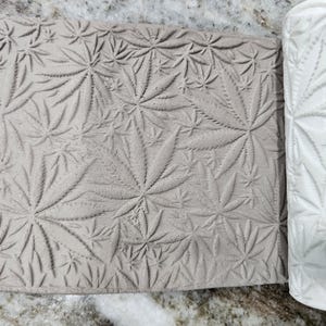 May include: A white cylindrical clay texture roller featuring a repeating embossed cannabis leaf pattern, shown next to a flat piece of light gray clay that has been imprinted with the same detailed botanical leaf design.