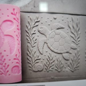 May include: A cylindrical pink silicone rolling pin with a recessed sea turtle, seaweed, and seashell pattern. Next to it is a flat piece of gray clay showing the embossed impression of the same sea turtle, seaweed, and seashell design.