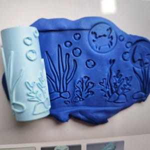May include: A light blue cylindrical rolling pin with an embossed underwater pattern featuring seaweed, bubbles, and a crab, shown next to a flat piece of dark blue modeling clay imprinted with the same detailed ocean-themed design.