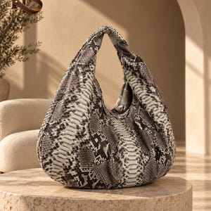 Peut inclure: Un sac à main de style hobo arborant un motif peau de serpent gris, noir et blanc. Le sac présente une silhouette souple et non structurée avec une poignée intégrée unique sur le dessus, formant une ouverture triangulaire. La texture imite les écailles.