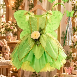 May include: A lime green fairy dress featuring attached sheer wings, a bodice with gold glitter accents, and a layered tulle skirt adorned with a large cream-colored flower at the waist. Includes a matching butterfly headband and a wand with streamers.