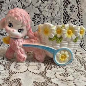 May include: A ceramic figurine of a pink poodle pulling a white cart shaped like a basket. The cart is decorated with yellow and white daisy flowers, and the wheel features a single daisy. The poodle has large black eyes and pink fur.