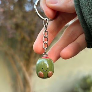 May include: A keychain featuring a small, smooth, olive-green charm shaped like a rounded olive. The charm has a hand-painted black smiley face, two small brown feet, and a red pimento top. It is attached to a silver-toned metal split ring.
