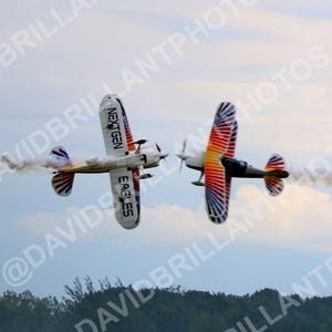 May include: Two aerobatic biplanes flying in formation against a cloudy sky. The left plane is white with the text NEXTGEN EAGLES on the wing. The right plane features a vibrant rainbow-colored wing pattern. Both aircraft emit trails of white smoke.