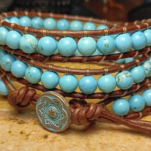 May include: Wrap bracelet with turquoise beads and brown leather strands. The bracelet has a button clasp with a turquoise and bronze finish. The turquoise beads are approximately 1 cm in diameter.