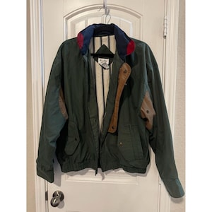 Puede incluir: Una chaqueta de color verde oscuro con un cuello multicolor en azul marino, rojo y verde oscuro. La chaqueta incluye detalles de cuero marrón en el pecho y las mangas, cierre de cremallera, bolsillos con botones a presión y forro a rayas crema.