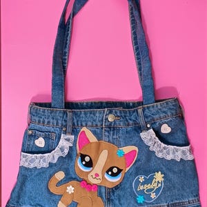 Op de afbeelding: Een blauwe denim tas met een bruine kat-applicatie met grote blauwe ogen, roze oren en een roze knoopkraag. De tas is voorzien van witte kanten randen op de zakken, een hartvormige patch met het woord Lovely en een geplooide onderrand.