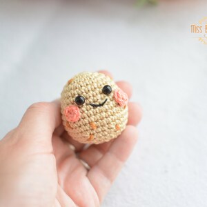 Può includere: Un uovo di colore beige lavorato a uncinetto con un viso sorridente, occhi neri e guance rosa.