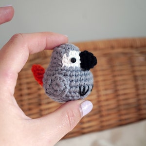 May include: A small gray crocheted bird with a black beak and white eyes. The bird has a red tail and is being held in a hand.
