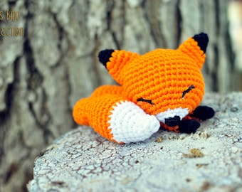 May include: A small orange and white crocheted fox toy with black eyes and a black nose, sleeping on a gray tree stump.