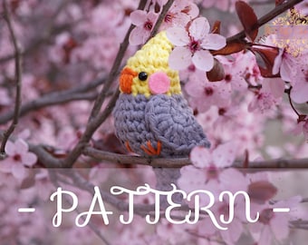 May include: A crocheted gray and yellow bird with orange accents perched on a branch of a pink flowering tree. The text "PATTERN" is written in white on a blurred background.
