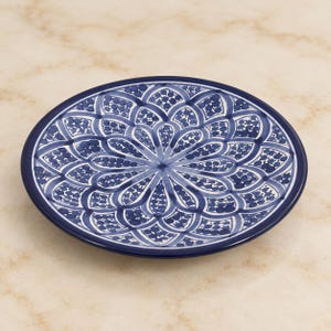 May include: A round ceramic plate featuring a dark blue and white hand-painted floral pattern. The design consists of symmetrical petal-like shapes radiating from the center, filled with small dots and intricate line work, finished with a solid blue rim.