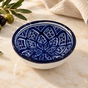 May include: A small, round ceramic bowl featuring a white exterior and a deep blue interior decorated with a hand-painted geometric pattern of diamonds and intricate floral-like dots. The rim is painted with a solid dark blue band.
