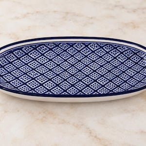 May include: An oval ceramic serving platter featuring a deep blue and white geometric diamond pattern across the interior surface. The rim is defined by a solid dark blue border, and the exterior base of the dish is a smooth, solid white ceramic.