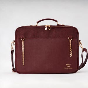 May include: A deep burgundy briefcase featuring a top handle, gold-tone zipper closures, and two decorative gold-tone chain accents on the front. The bag includes a detachable shoulder strap and the gold-colored text VELORAWORK printed on the lower right.