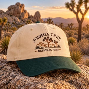 May include: A cream-coloured baseball cap with a dark green brim. The front features a brown embroidered graphic of a desert landscape with Joshua trees and the text JOSHUA TREE NATIONAL PARK in a curved, dark brown font.