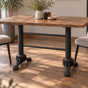 Puede incluir: Una mesa de comedor rectangular con una superficie de grano de madera y una base de metal negro con dos columnas verticales conectadas por barras horizontales, que terminan en pies decorativos en forma de garra.