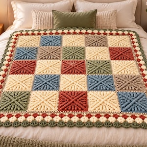 May include: A square crochet blanket featuring a grid of textured squares in cream, sage green, dusty blue, and terracotta. The blanket is finished with a decorative scalloped border composed of small cream flowers and green leaf-like edges.