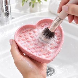 Op de afbeelding: Een hartvormige roze siliconen make-up kwastenreinigingsmat met kleine verhoogde hartjes op het oppervlak. De mat is bedekt met witte zeepbellen en wordt gebruikt om een make-up kwast met donkere haren en een licht handvat te reinigen.
