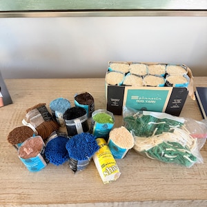 Könnte beinhalten: Eine Sammlung von Spinnerin Teppichgarn, bestehend aus einem Karton mit acht cremefarbenen Garnknäueln, mehreren einzelnen Knäueln in Braun, Blau, Schwarz, Grün und Rosa sowie einem Plastikbeutel mit losen grünen und cremefarbenen Garnsträngen.