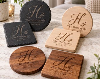 Puede incluir: Un conjunto de seis posavasos redondos y cuadrados, hechos de pizarra oscura, madera clara y madera oscura. Cada pieza está grabada con una letra H grande, el texto The Hufnagels y Est. 2026, junto con un pequeño diseño de rama de hojas.