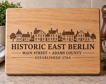 Puede incluir: Una tabla de cortar rectangular de madera marrón claro con un grabado láser marrón oscuro de una hilera de casas históricas. El texto dice: HISTORIC EAST BERLIN, MAIN STREET, ADAMS COUNTY, ESTABLISHED 1764, centrado en la tabla.