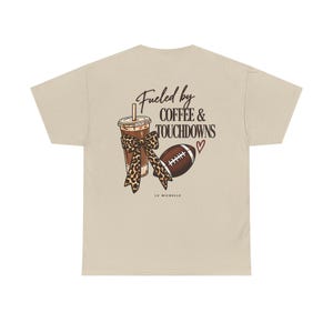 Könnte beinhalten: Ein beiges T-Shirt mit einem Grafikdruck auf der Rückseite und dem Text "Fueled by COFFEE & TOUCHDOWNS". Das Motiv zeigt einen Eiskaffee mit einer Schleife im Leopardenmuster, einen braunen Football und ein kleines rotes Herz. "LU MICHELLE" steht darunter.