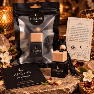 May include: A black car diffuser with a light wood cap and a black cord, packaged in a clear plastic pouch with a black label featuring the text HELLOIR. Includes a black card with gold text and a white card with printed inspirational text.