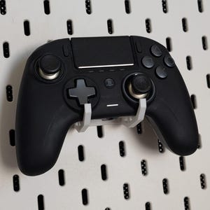 Könnte beinhalten: Ein schwarzer kabelloser Videospiel-Controller ruht auf einer weißen Wandhalterung aus Kunststoff. Der Controller verfügt über ein zentrales Touchpad, ein Steuerkreuz, zwei Analogsticks mit silbernen Basen und vier Aktionstasten auf der rechten Seite.