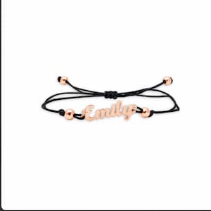 May include: A black cord bracelet featuring a rose gold-toned metal nameplate with the name Emily in a cursive script font. The bracelet includes two small rose gold-toned beads on either side of the nameplate and two at the ends of the cord.