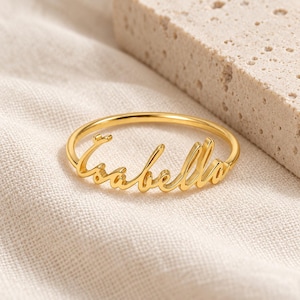 May include: A gold-toned ring featuring the name Isabella written in a cursive script font. The ring has a thin, polished band that connects to the lettering, which is integrated into the design. The metal surface reflects light with a smooth finish.