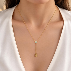 May include: A gold-toned lariat necklace featuring a delicate chain, a single round light blue gemstone set in a bezel, and a dangling capital letter R pendant. The necklace rests against a white garment, showcasing a minimalist design.