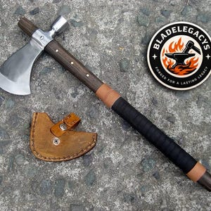 May include: A metal tomahawk with a dark wood handle, black grip, and tan leather accents, accompanied by a tan leather blade sheath. A circular sticker features the text BLADELEGACYS FORGED FOR A LASTING LEGACY with an anvil and fire graphic.