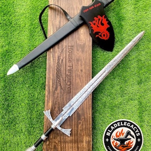May include: A metal sword with a patterned blade and a black handle, accompanied by a black scabbard featuring a red three-headed dragon emblem and the text Fire and Blood. The set rests on a wooden plaque engraved with House Targaryen.