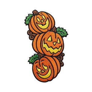 Puede incluir: Un parche bordado que muestra una pila vertical de tres calabazas naranjas con caras talladas en amarillo y hojas verdes. Cada calabaza tiene una expresión sonriente con ojos triangulares y una boca dentada, delineada en hilo marrón.