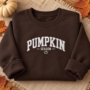 Op de afbeelding: Een donkerbruin sweatshirt met ronde hals en witte geborduurde tekst die PUMPKIN SEASON leest in een gebogen lettertype, met een klein wit geborduurd pompoenpictogram direct onder de tekst op de voorzijde van het kledingstuk.