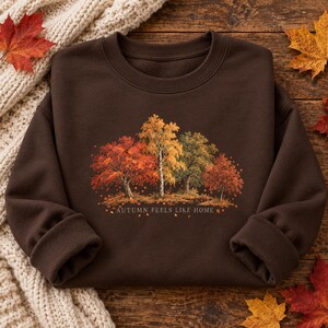 May include: A dark brown crewneck sweatshirt featuring a central graphic of four trees with autumn foliage in shades of red, orange, and yellow. Below the trees, the text reads "AUTUMN FEELS LIKE HOME" in a simple, light-colored font.