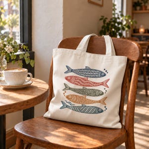 May include: A cream-colored canvas tote bag featuring a vertical graphic print of five stylized fish stacked on top of each other. The fish are illustrated in blue, red, green, and orange with intricate geometric patterns and small circular bubbles.