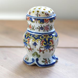 Könnte beinhalten: Ein Keramikstreuer mit cremefarbener Basis und handgemaltem Blumenmuster in Blau, Gelb, Grün und Rot. Der Streuer hat einen abgerundeten Körper, einen ausgestellten Fuß und einen perforierten Deckel mit mehreren Löchern zum Dosieren.