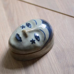 Op de afbeelding: Een klein, ovaalvormig keramieken doosje met een deksel waarop een gebeeldhouwd menselijk gezicht is afgebeeld. Het gezicht is geglazuurd in lichtgrijs met donkerblauwe details, waaronder ogen, lippen, stervormige markeringen op de wangen en haar.